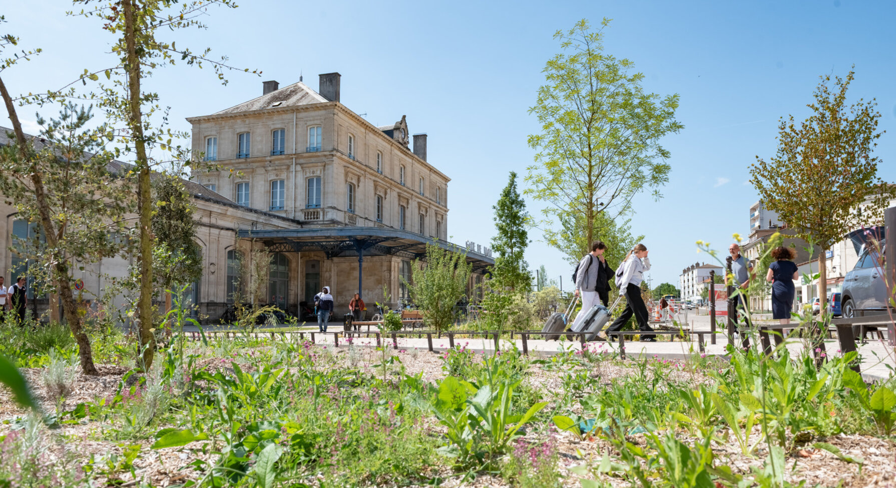 Esplanade réaménagée devant la gare de Niort avec jeunes arbres et plantations, des voyageurs avec bagages circulent