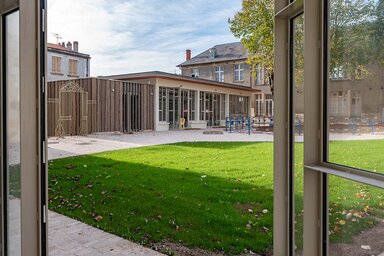 Chantier en fin de réhabilitation : pelouse et pavillon en bois neuf encadrés de bâtiments anciens, vus depuis une baie vitrée ouverte.
