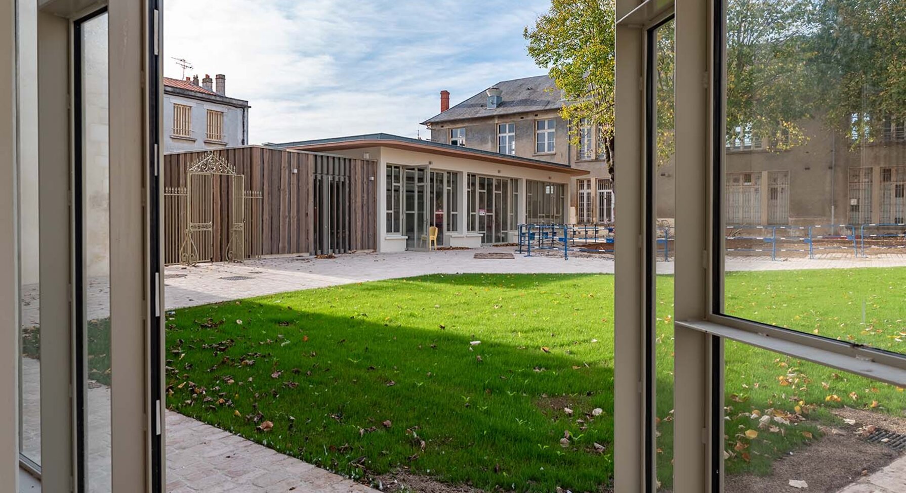 Chantier en fin de réhabilitation : pelouse et pavillon en bois neuf encadrés de bâtiments anciens, vus depuis une baie vitrée ouverte.